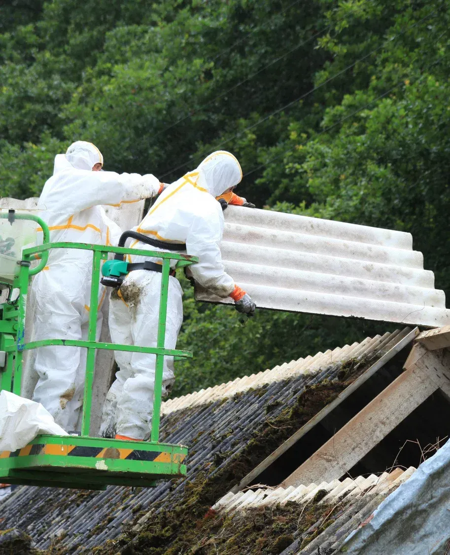 Two workers in PPE removing corrugated asbestos roofing from a building.