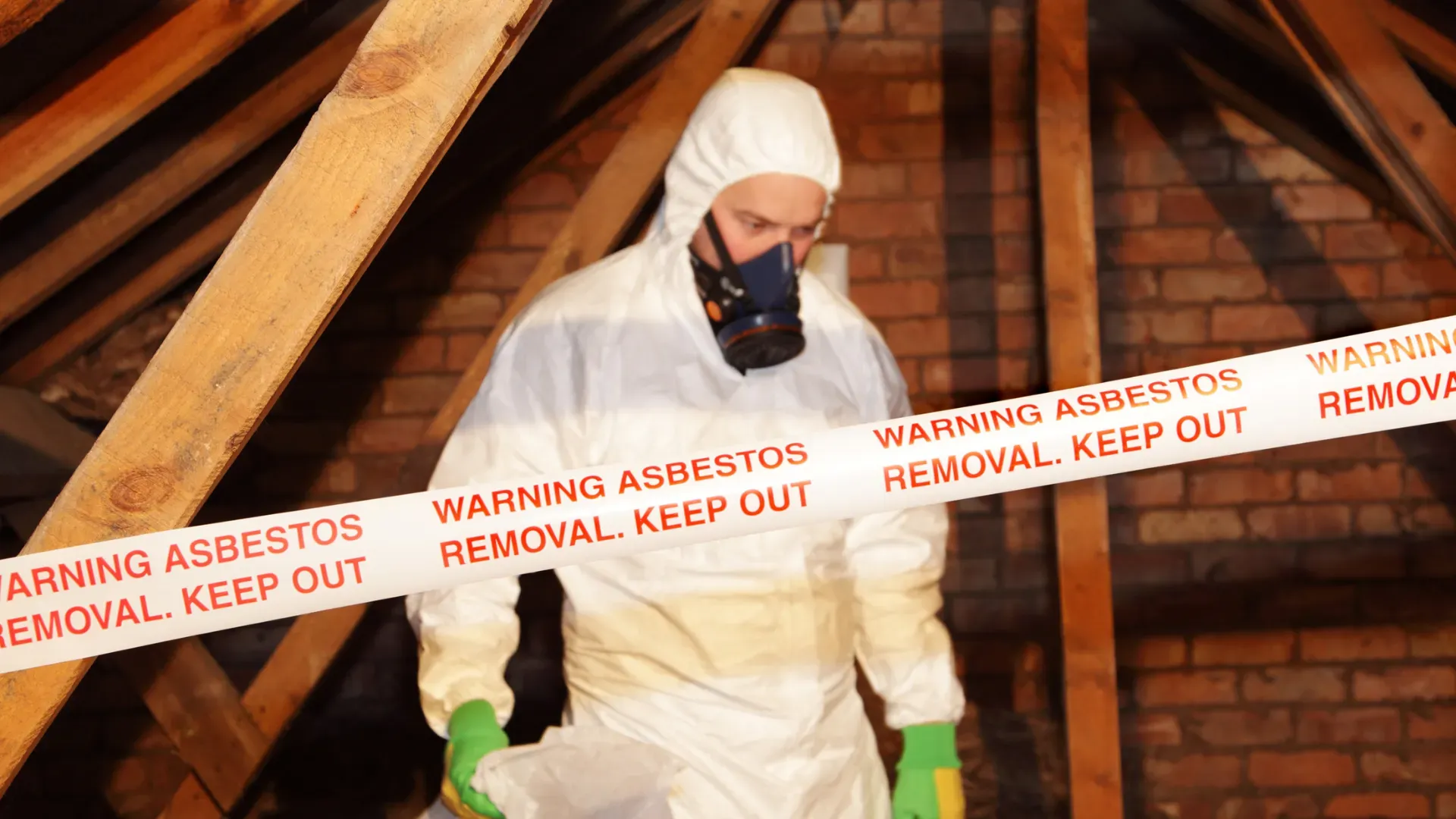 Worker in protective suit with mask and gloves, standing in an attic with warning tape indicating asbestos removal. The tape says 'WARNING ASBESTOS REMOVAL. KEEP OUT'.
