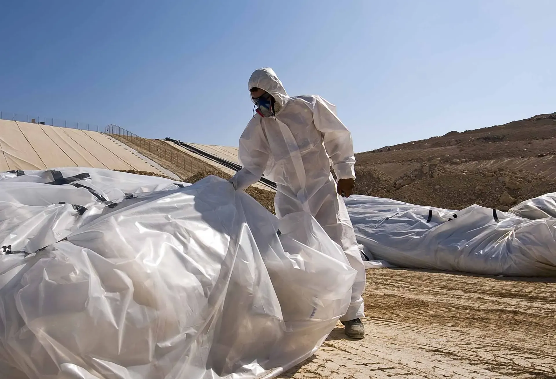 Worker in protective suit and mask handling large white bags of waste outdoors.