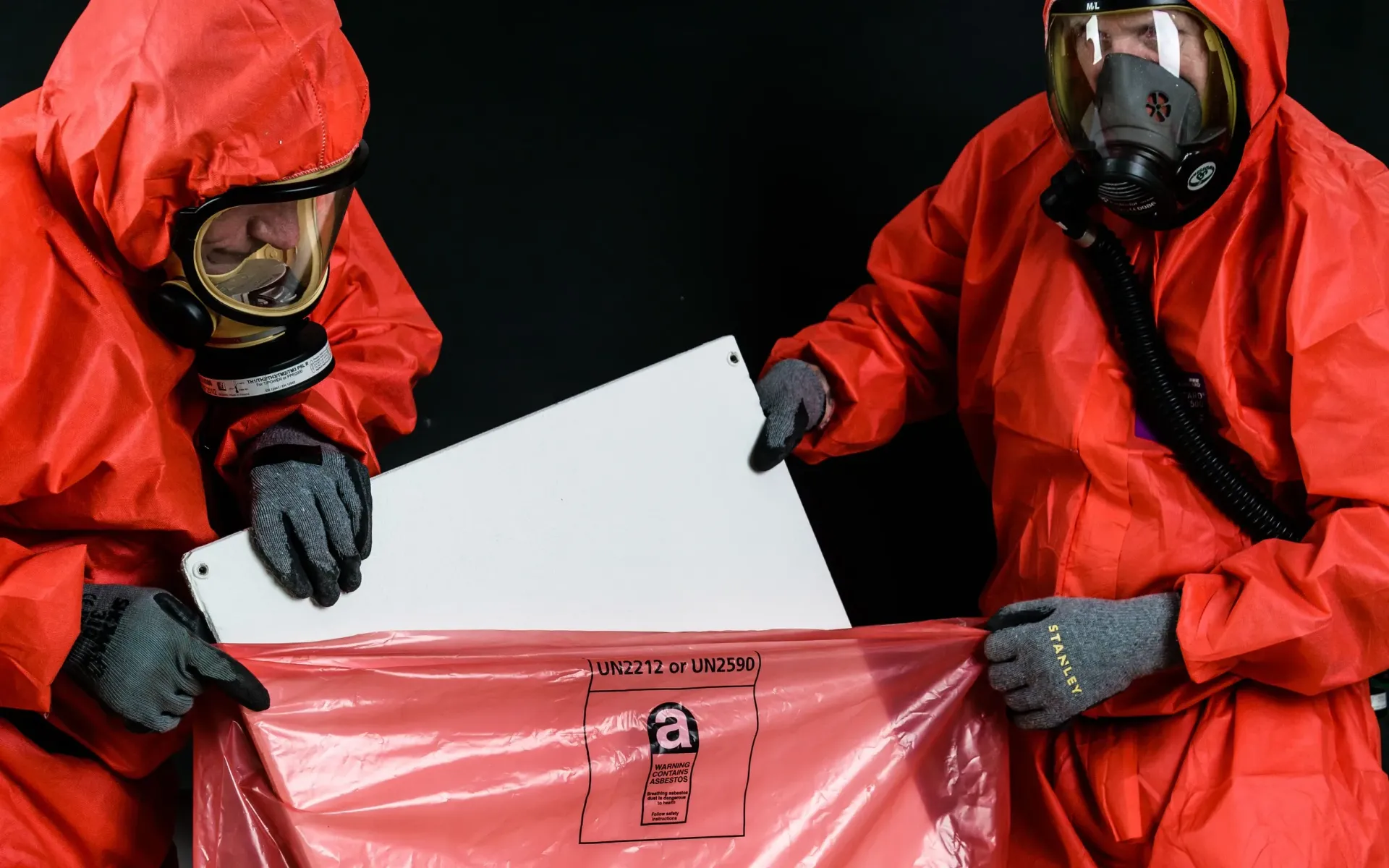 Asbestos professionals in PPE depositing asbestos containing material into a sealed waste bag.
